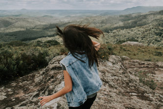 Girl in blue sleeveless denim jacket stands on top of mountain with brown hair blowing in wind and arms out to her sides