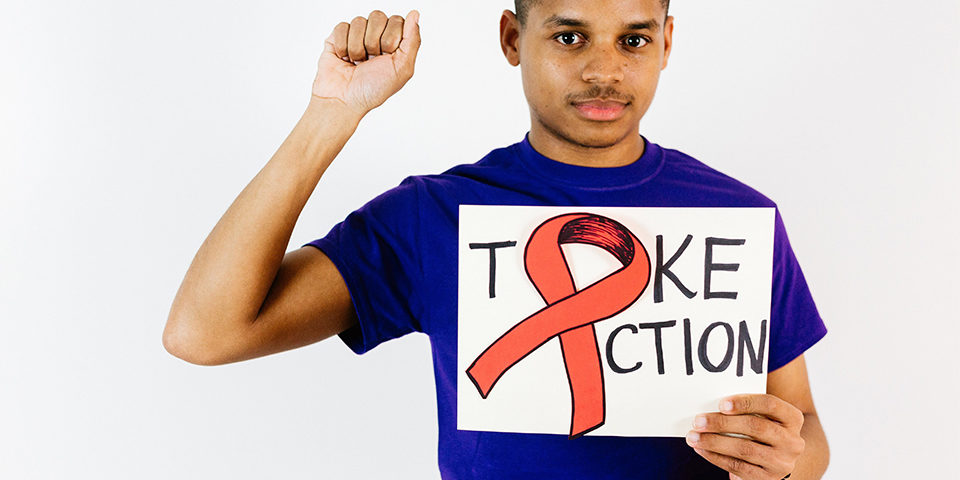 young man in indigo shirt holds sign with red ribbon that says "take action" while holding up right fist