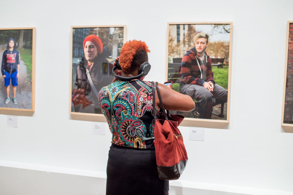 Woman in patterned top with red handbag and red-orange mohawk listens to audio through over-the-ear headphones while looking at row of portraits.