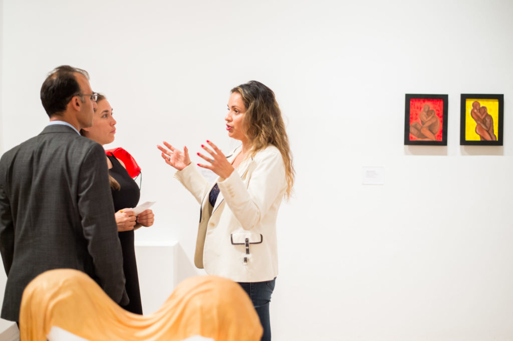 Woman in white blazer gestures with both hands while speaking to woman in black dress and man in gray plaid suit jacket and glasses in art gallery.