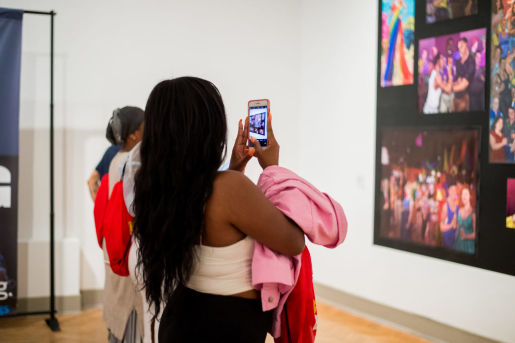 Girl in white tank top carrying a pink jacket taking photo of large art piece on wall