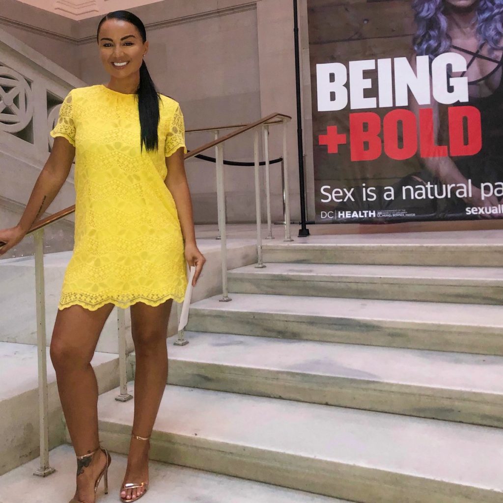 Women in yellow lace dress and metallic heels posing on staircase; Banner in back with girl with purple hair and "BEING + BOLD" 