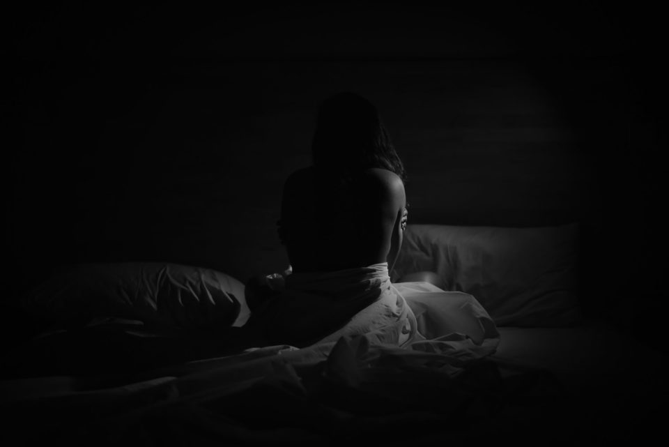 silhouette of young woman sitting on bed, holding her arms and looking away from the viewer.