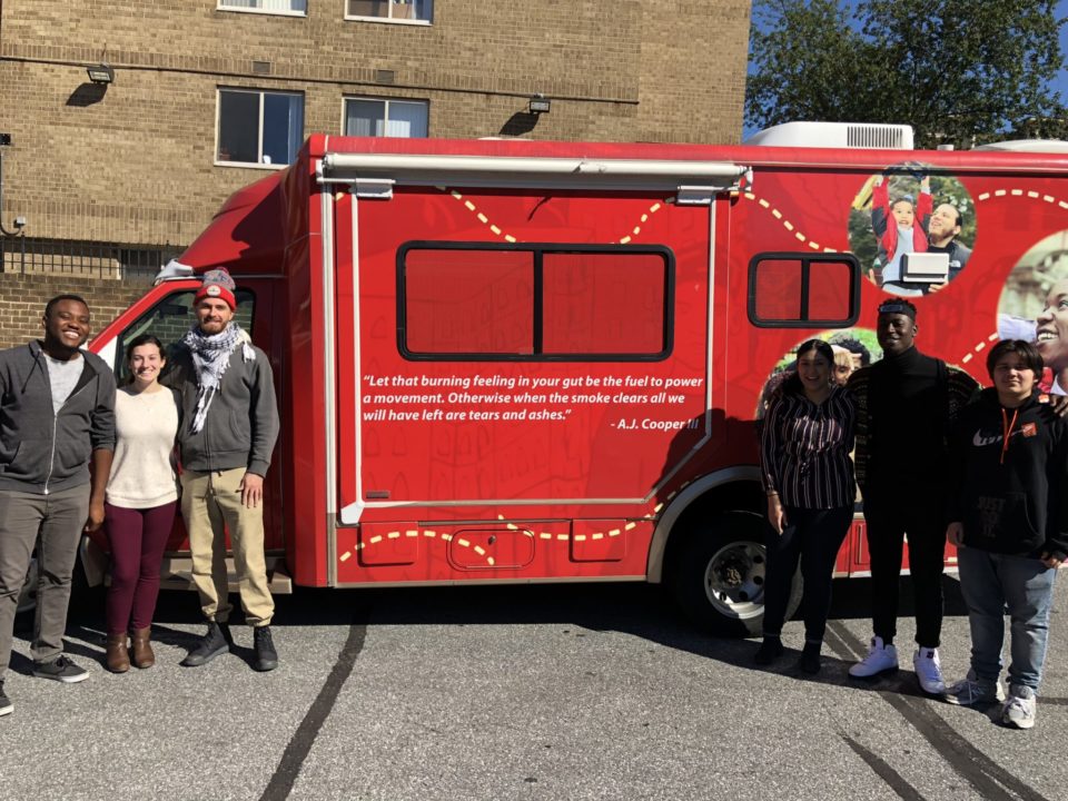 Member of the Latin American Youth Center stand in front of a red vehicle with the quote "Let that burning feeling in your guy be the fuel to power a movement. Otherwise when the smoke clears all we will have left are tears and ashes" - A.J. Cooper