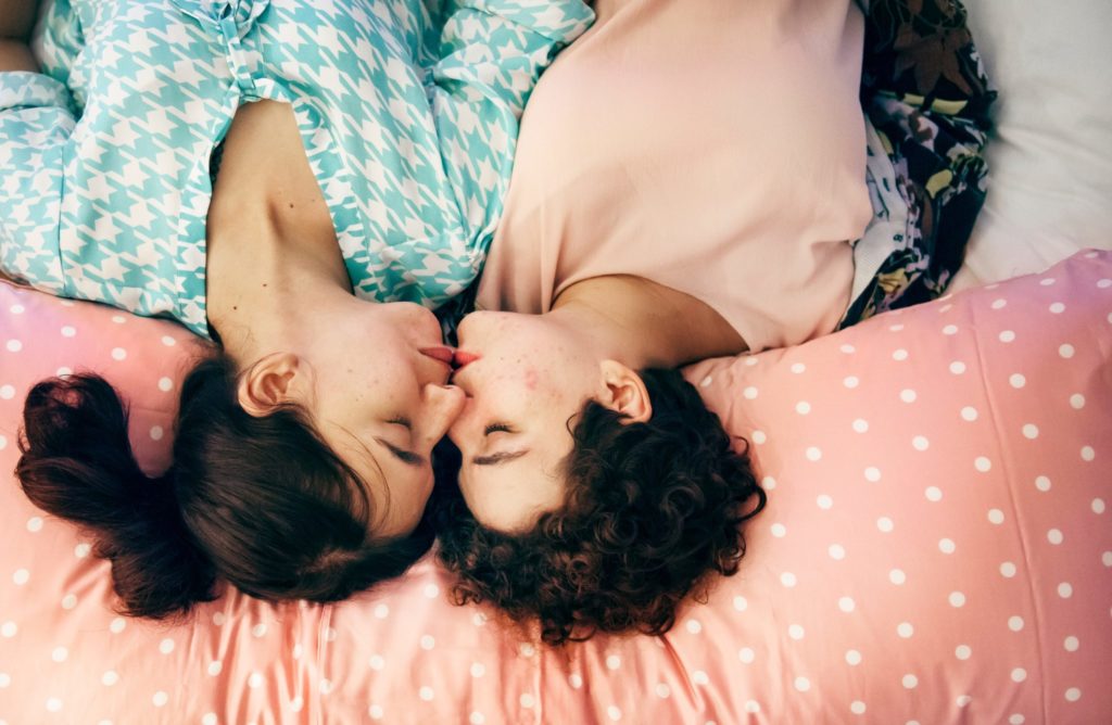 two women lie on pink comforter and slightly kiss