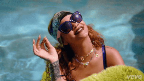 Rihanna chilling in a pool with green fur around her, sunglasses, and a patterned scarf on her head waving to the camera.