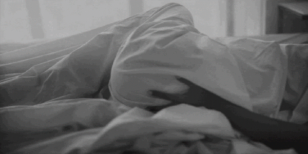 Woman laying in bed under sheets rubbing hand up lower back. Image is in Black and White.